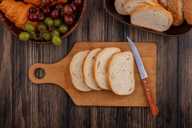 Bovenaanzicht van brood als gesneden gezaaide bruine cob en witte in kom en op snijplank met mes en mandje van croissant druif op houten achtergrond