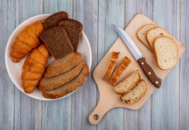 Bovenaanzicht van brood als gesneden stokbrood met mes op snijplank en croissantrogge en gezaaide bruine cob in kom op houten achtergrond