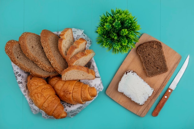 Bovenaanzicht van brood als gesneden stokbrood met zaadjes en croissant in kom en roggebrood besmeurd met kaas op snijplank met mes en plant op blauwe achtergrond