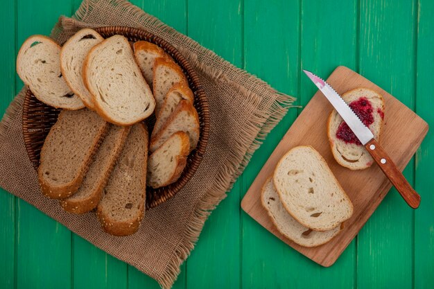 Bovenaanzicht van brood als gezaaide bruine cob en stokbrood plakjes in mand op zak en sneetje brood besmeurd met frambozenjam met mes op snijplank op groene achtergrond