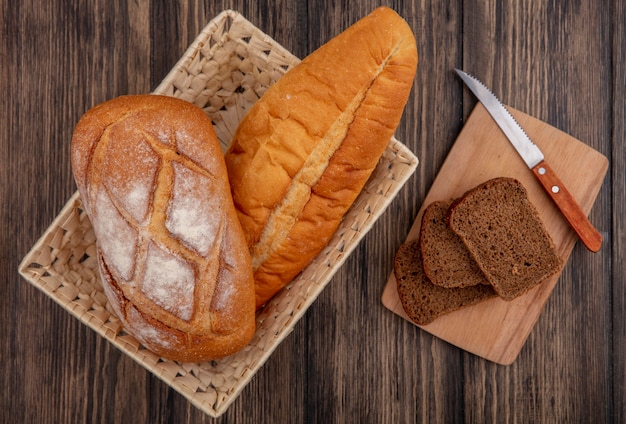 Bovenaanzicht van brood als knapperig en Vietnamees stokbrood in mand en gesneden roggebrood met mes op snijplank op houten achtergrond