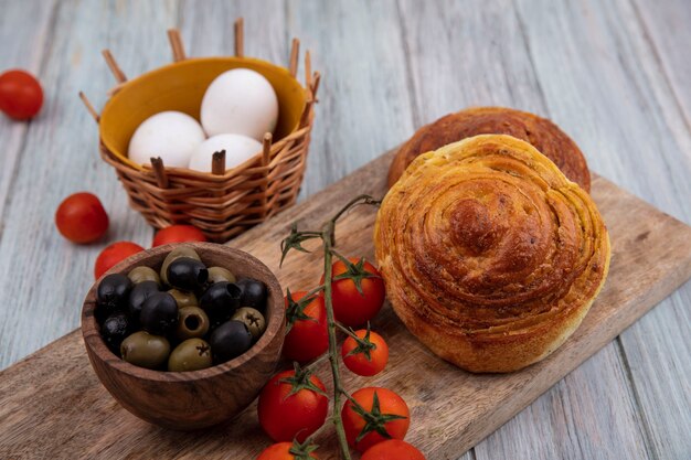 Bovenaanzicht van broodjes op een houten keukenbord met verse trostomaten met olijven op een houten kom en eieren op een emmer op een grijze houten achtergrond