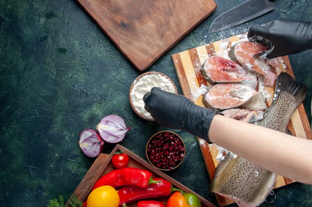 Bovenaanzicht van chef-kok die rauwe visplakken bedekt met meel, verse groenten op houten bord, meelkommes op keukentafel