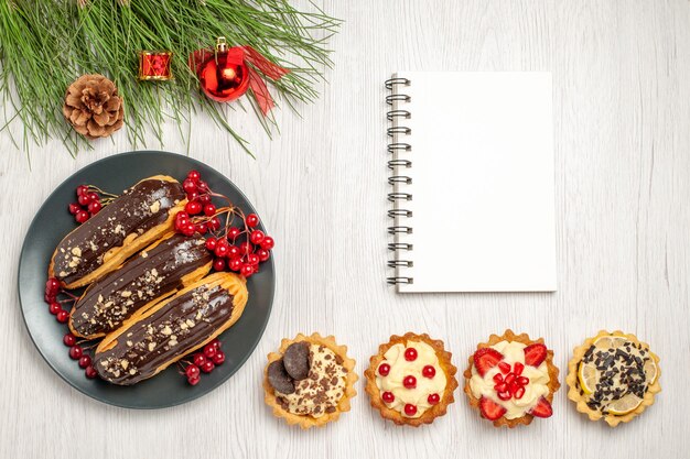 Bovenaanzicht van chocolade-eclairs en aalbessen op de grijze plaat een notebook-taartjes aan de onderkant en dennenboom bladeren met kerstspeelgoed op de witte houten grond