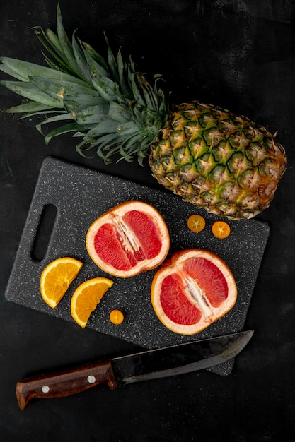 Bovenaanzicht van citrusvruchten als grapefruit oranje ananas en kumquat met mes op snijplank op zwarte ondergrond