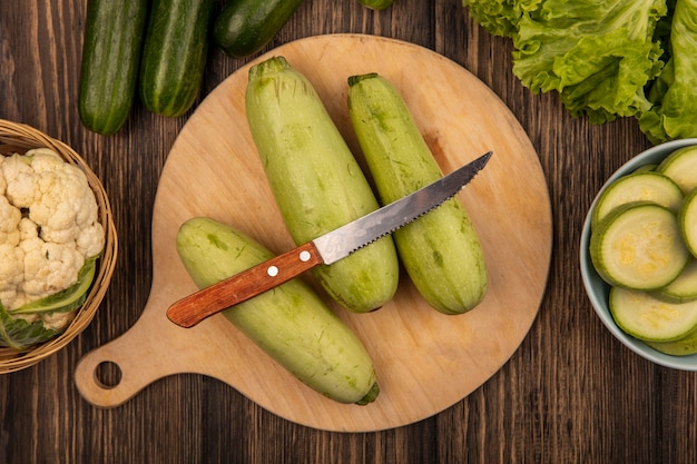 Bovenaanzicht van courgettes geïsoleerd op een houten keukenbord met mes met bloemkool op een emmer met komkommers en sla geïsoleerd op een houten oppervlak