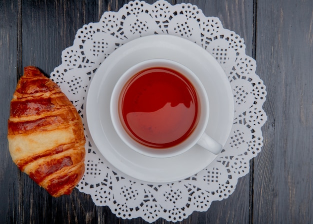 Gratis foto bovenaanzicht van croissant en kopje thee op theezakje op papier kleedje en houten achtergrond