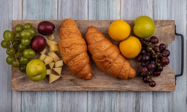 Bovenaanzicht van croissants met druiven, plukken nectacots en kaas op snijplank op houten achtergrond