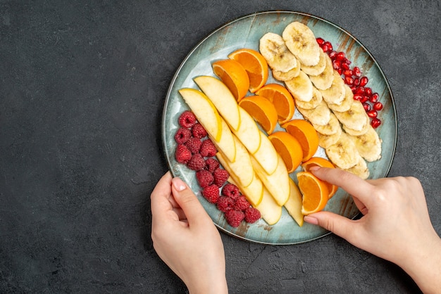 Bovenaanzicht van de hand die stukjes sinaasappel neemt, het verzamelen van gehakt vers fruit op een blauw bord op zwarte tafel