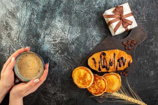 Bovenaanzicht van de hand met een kopje koffie en een smakelijk ontbijt met pannenkoeken croisasant en geschenkdoos op donkere tafel