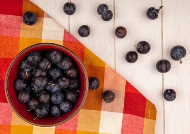 Bovenaanzicht van de kleine zure blauw-zwarte fruit sleepruimen op een rode kom met sleepruimen geïsoleerd op een witte houten achtergrond