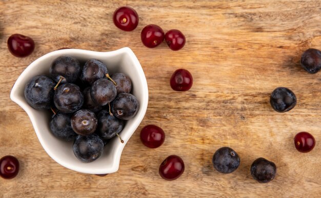 Bovenaanzicht van de kleine zure blauwzwarte fruit sleeën op een witte kom met rode kersen geïsoleerd op een houten achtergrond