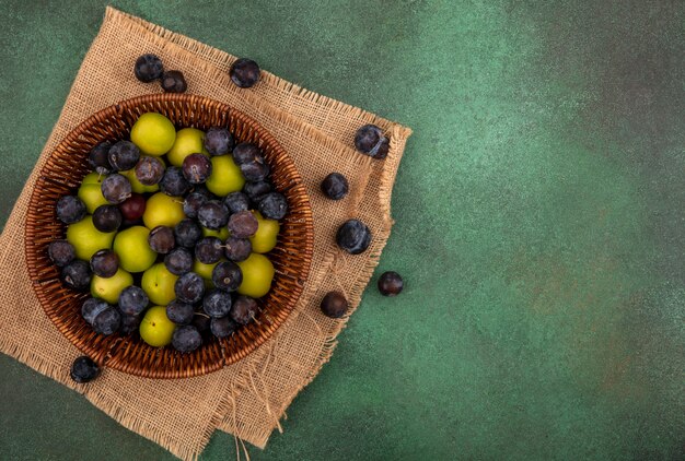 Bovenaanzicht van de kleine zure blauwzwarte fruit sleepruimen met groene kersenpruim op een emmer op een zakdoek op een groene achtergrond met kopie ruimte