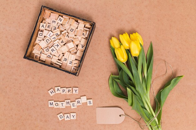 Bovenaanzicht van de tekst van de gelukkige moederdag; gele tulp bloemen; prijskaartje en houten blokken over bruine achtergrond