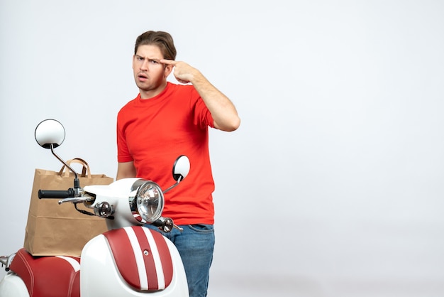 Bovenaanzicht van denken bezorger in rood uniform staande in de buurt van de bestelling van de scooter op witte muur