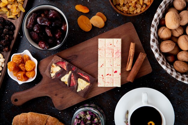 Bovenaanzicht van donkere en witte chocoladerepen met gedroogde vruchten op een houten bord, kaneelstokjes, walnoten en een kopje thee op rustiek