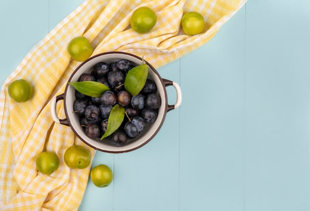 Bovenaanzicht van donkerpaarse sleepruimen op een kom met groene kersenpruimen geïsoleerd op een geel geruit tafelkleed op een blauwe achtergrond met kopie ruimte