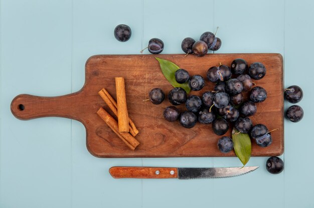 Bovenaanzicht van donkerpaarse zure sleepruimen op een houten keukenbord met kaneelstokjes met mes op een blauwe achtergrond