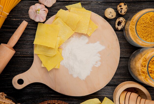 Bovenaanzicht van dun gerold rauw deeg in vierkanten gesneden op een houten bord met bloem en verschillende soorten Italiaanse rauwe pasta in glazen potten op zwarte houten achtergrond