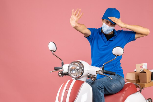 Bovenaanzicht van een bezorgde bezorger met een medisch masker met een hoed die op een scooter zit en een stopgebaar maakt op pastel perzik