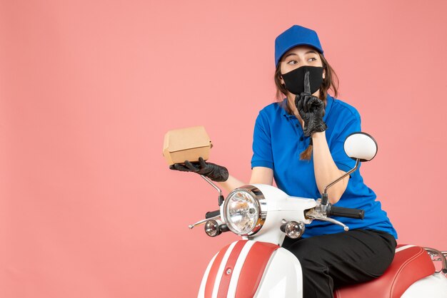 Bovenaanzicht van een bezorger met een medisch masker en handschoenen die op een scooter zit en bestellingen aflevert die een stiltegebaar maken op een pastelkleurige perzikachtergrond