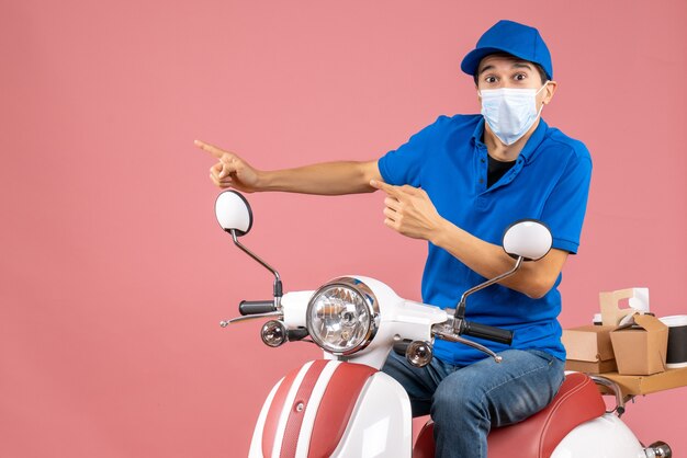 Bovenaanzicht van een bezorger met een medisch masker met een hoed die op een scooter zit en iets aan de rechterkant wijst op een pastelkleurige perzikachtergrond