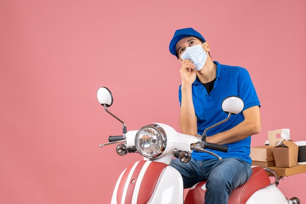 Bovenaanzicht van een denkende koeriersman met een medisch masker met een hoed die op een scooter zit en bestellingen aflevert op pastel perzik