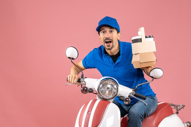 Bovenaanzicht van een geschokte koeriersman met een medisch masker met een hoed die op een scooter zit en de bestelling vasthoudt op een pastelkleurige perzikachtergrond