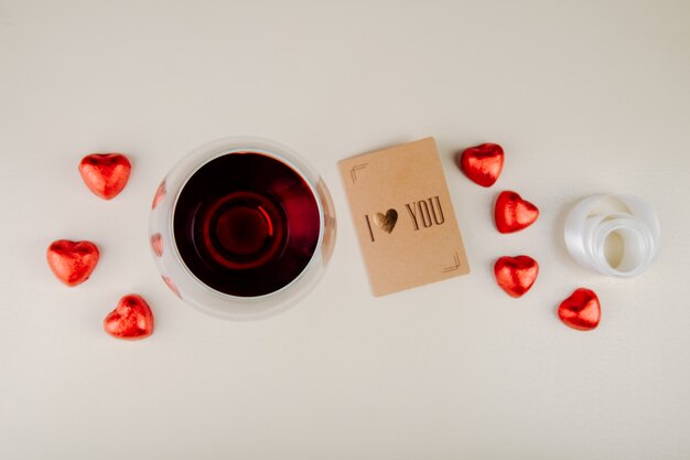 Bovenaanzicht van een glas wijn met hartvormige chocoladesuikergoed verpakt in rode folie en een kleine ansichtkaart op witte tafel