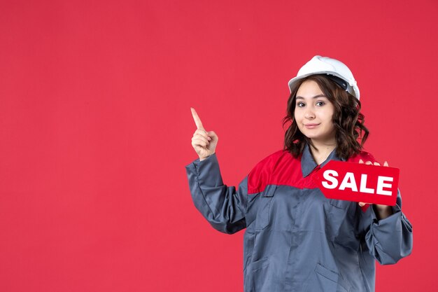 Bovenaanzicht van een glimlachende vrouwelijke bouwer in uniform met een helm en een verkooppictogram dat aan de rechterkant op geïsoleerde rode achtergrond omhoog wijst