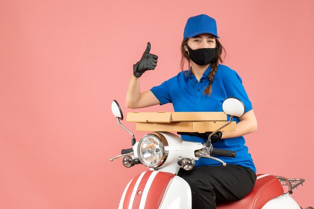 Bovenaanzicht van een glimlachende vrouwelijke koerier met een medisch masker en handschoenen die op een scooter zitten en bestellingen afleveren die een goed gebaar maken op pastel perzik