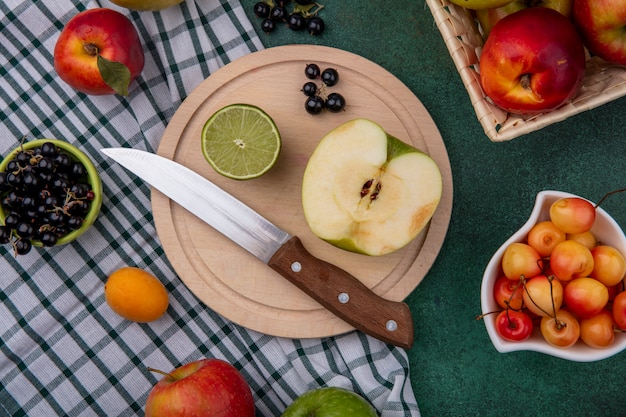 Bovenaanzicht van een halve groene appel met een mes op een stand met limoen, witte kersen, zwarte bessen en perziken op een geruite handdoek op een groen oppervlak