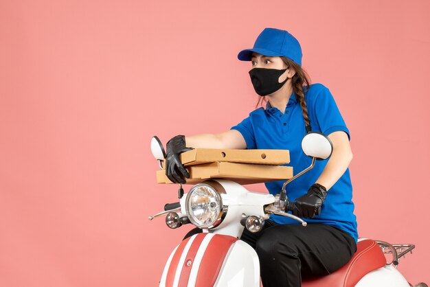 Bovenaanzicht van een jonge, nieuwsgierige, emotionele vrouwelijke koerier met een medisch masker en handschoenen die op een scooter zit en bestellingen aflevert op pastel perzik