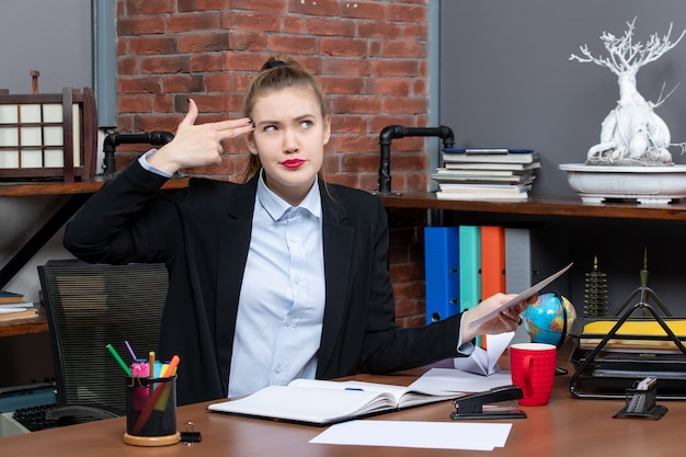 Gratis foto bovenaanzicht van een jonge vrouw die aan een tafel zit en een document vasthoudt en een pistoolgebaar maakt op kantoor