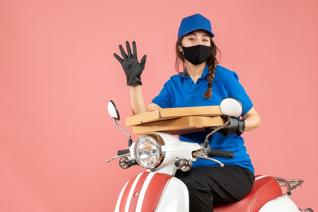 Bovenaanzicht van een jonge vrouwelijke koerier met een medisch masker en handschoenen die op een scooter zitten en bestellingen afleveren met vijf op pastel perzik