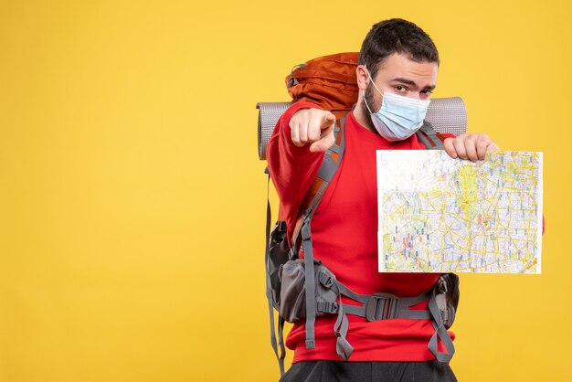 Bovenaanzicht van een jonge zelfverzekerde reiziger die een medisch masker draagt met een rugzak met een kaart die naar voren wijst op geel on