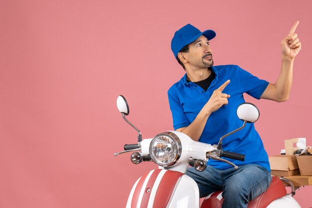 Bovenaanzicht van een koeriersman met een hoed die op een scooter zit en bestellingen aflevert die naar boven wijzen op een pastelkleurige perzikachtergrond