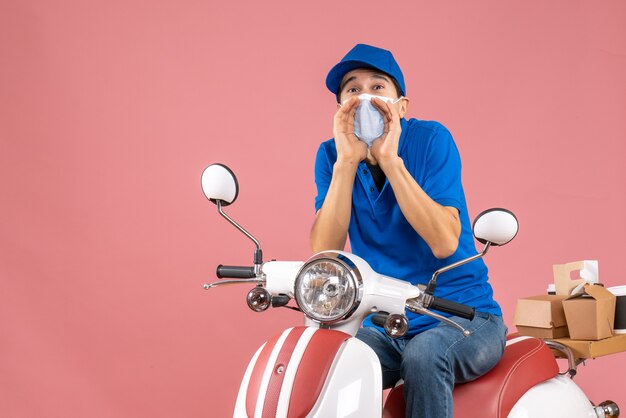 Bovenaanzicht van een koeriersman met een medisch masker met een hoed die op een scooter zit en iemand belt op een pastelkleurige perzikachtergrond