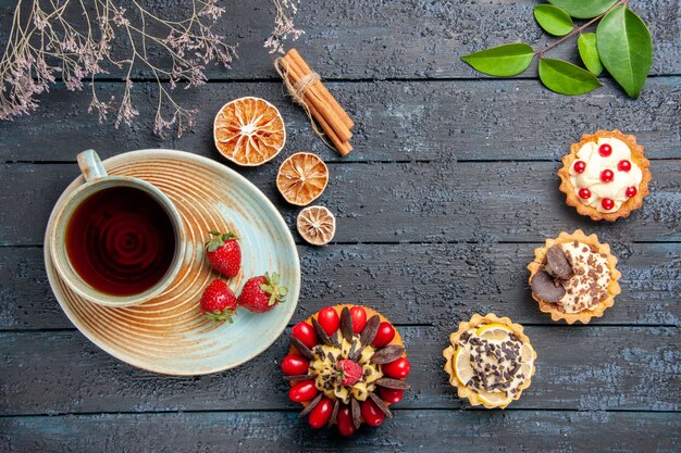 Bovenaanzicht van een kopje thee en aardbeien op schotel, gedroogde sinaasappeltaartjes, bladeren kaneel en bessencake op donkere houten tafel
