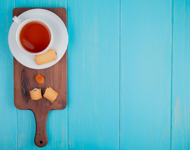 Bovenaanzicht van een kopje thee met koekjes en gedroogde abrikoos op houten snijplank op blauw met kopie ruimte