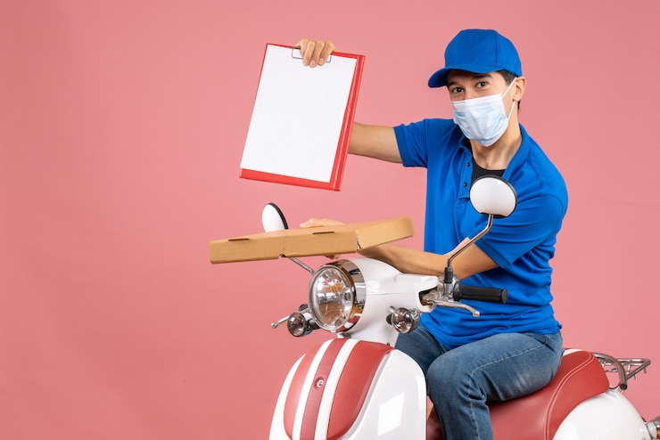 Gratis Foto bovenaanzicht van een mannelijke bezorger met een masker met een hoed die op een scooter zit en bestellingen aflevert met een document op een perzikachtergrond