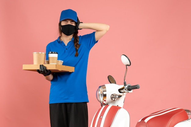 Bovenaanzicht van een nerveus koeriersmeisje met medische maskerhandschoenen die naast een motorfiets staan met koffiekoekjes op een pastelkleurige perzikkleurige achtergrond