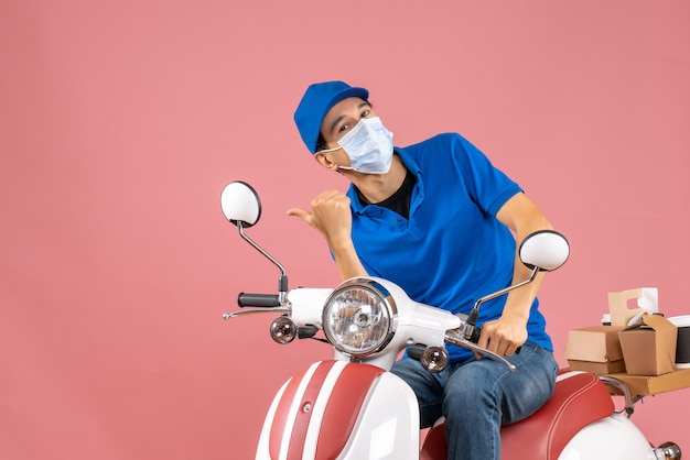 Bovenaanzicht van een nieuwsgierige koeriersman met een medisch masker met een hoed die op een scooter zit en iets wijst op een pastelkleurige perzikachtergrond