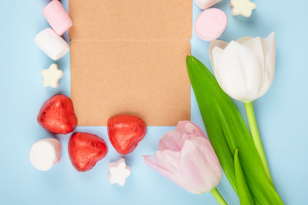 Bovenaanzicht van een open bruine papieren wenskaart met verspreide marshmallows en hartvormige chocoladesuikergoed in rode folie met roze kleurentulpen op blauwe tafel