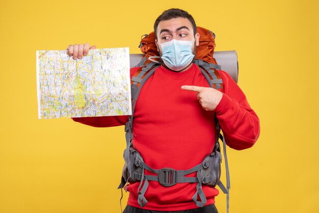 Bovenaanzicht van een reiziger die een medisch masker draagt met een rugzak die op een gele achtergrond wijst