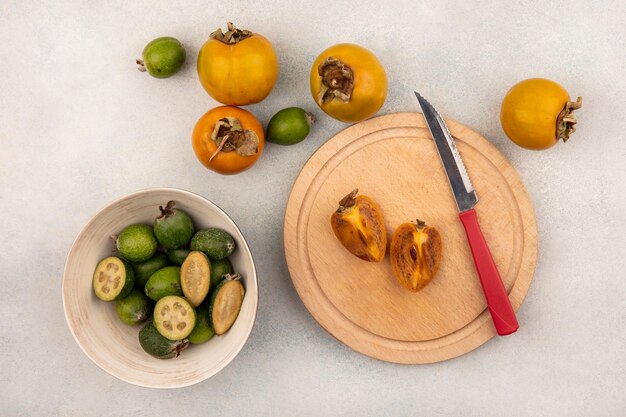 Bovenaanzicht van een rijpe halve kaki fruit op een houten keukenplank met mes met feijoas op een kom en kaki geïsoleerd op een grijze ondergrond