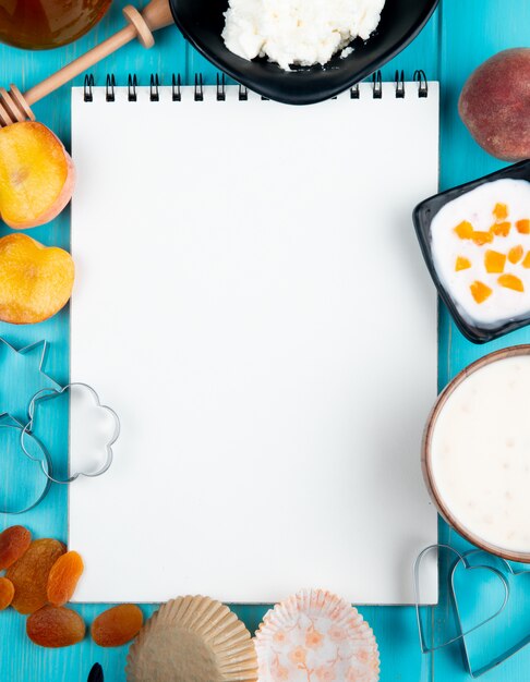 Bovenaanzicht van een schetsboek en verse rijpe perziken gedroogde abrikozen kwark yoghurt en cookie cutters gerangschikt op blauw