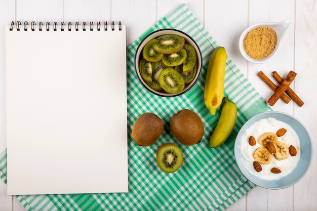 Bovenaanzicht van een schetsboek, gesneden kiwi in een kom, vers bananenfruit, yoghurt met gesneden banaan met amandel en kaneelstokjes op wit hout