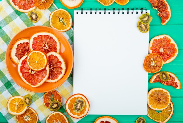 Bovenaanzicht van een schetsboek met gedroogde sinaasappel en grapefruit segmenten op een plaat en gedroogde kiwi op groene houten achtergrond