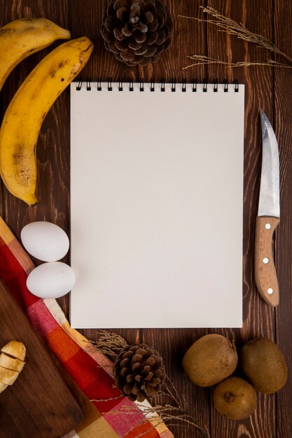 Bovenaanzicht van een schetsboek met kiwi en banaan fruit en twee eieren met keukenmes op hout